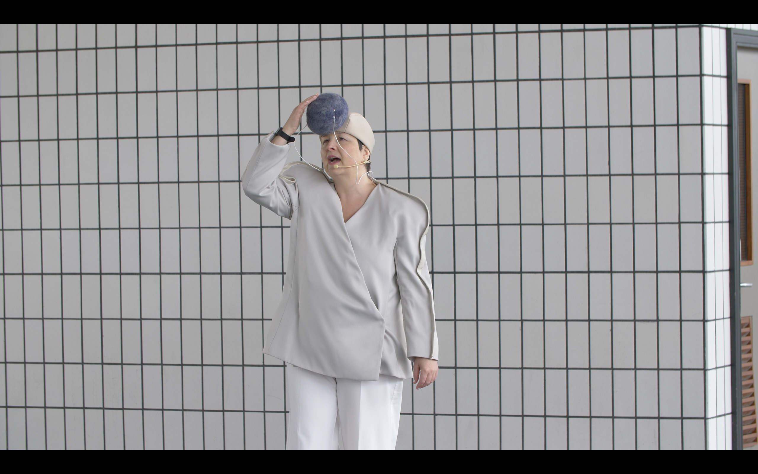 Image taken from a performance at the Bus Station, the artist is dressed in a white sound suit touching a large grey pom pom on their hat.