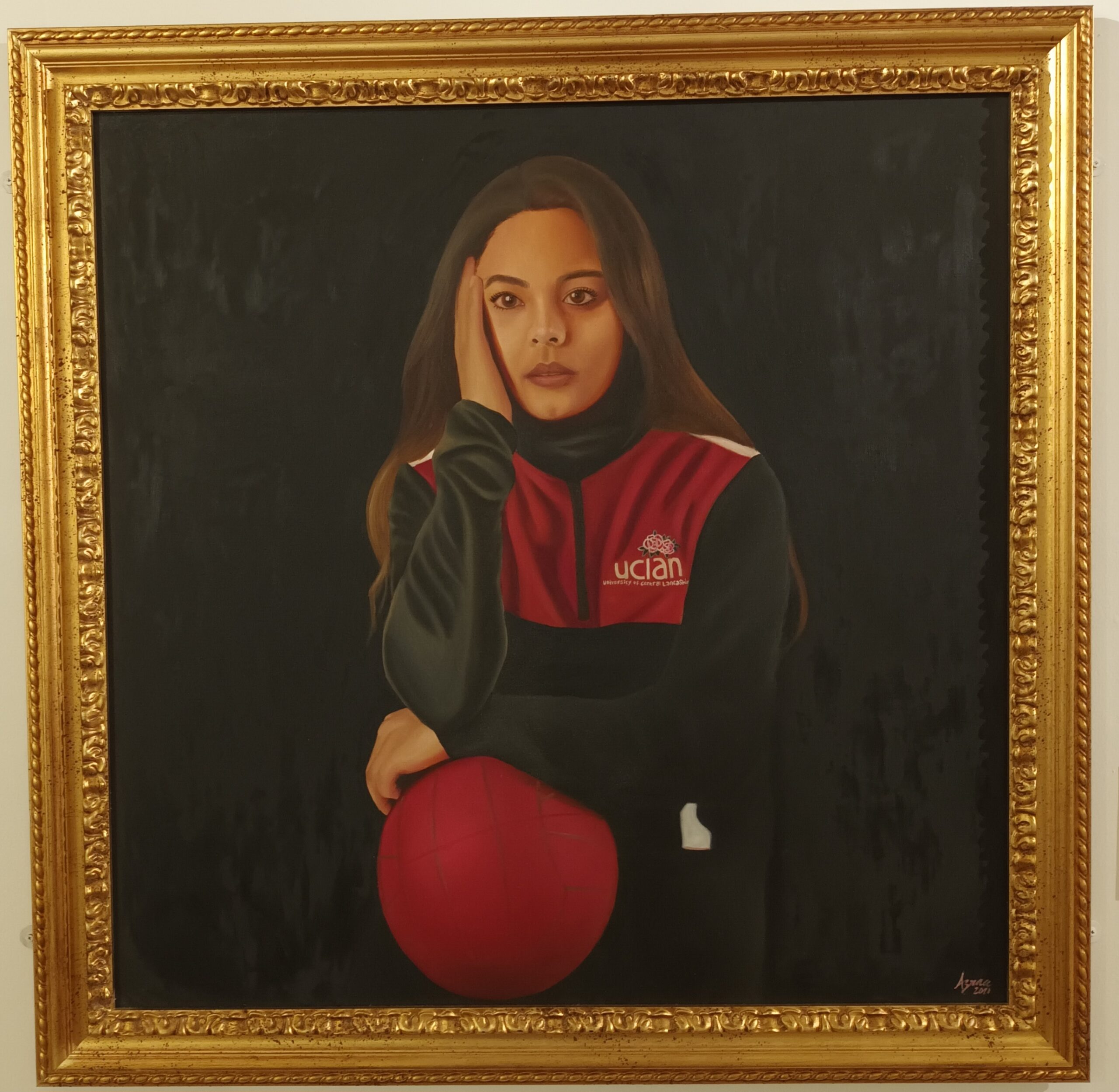 Image of a painting framed in gold of a person leaning on a ball wearing a UCLAN branded jacket.