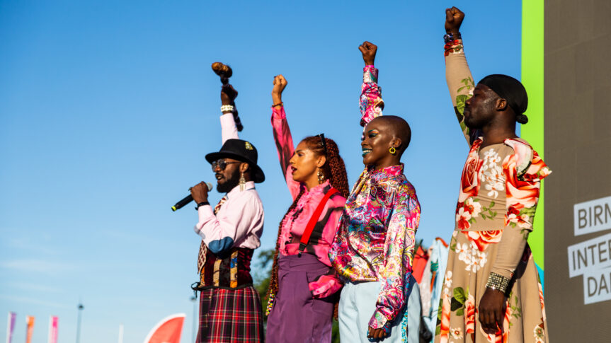 Four people dressed in colourful clothing stand side by side with their fists raised to the sky.