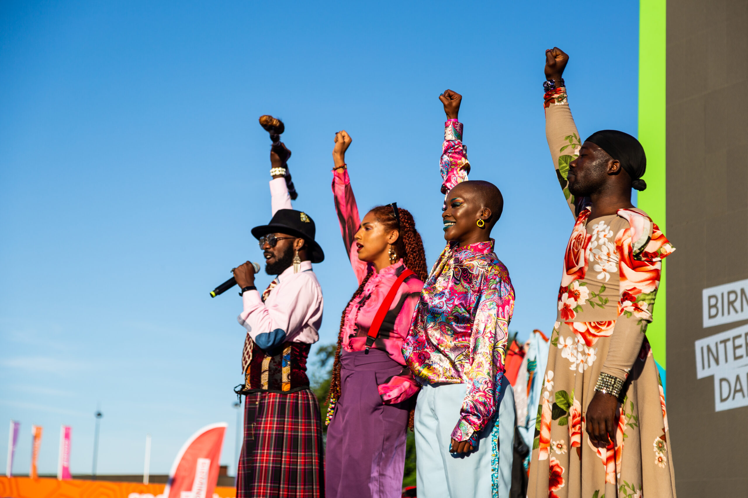 Four people dressed in colourful clothing stand side by side with their fists raised to the sky.