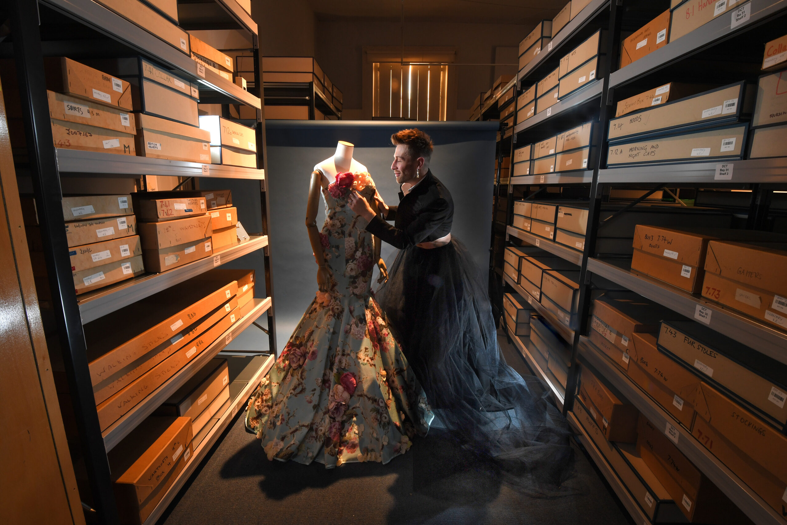 A person wearing a cropped jacket and long skirt stands next to a blue rose dress.