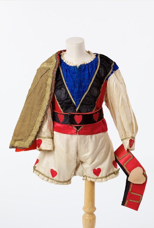 Knave of Hearts costume, red, white blue and black shirt, gold cloak with red lining. Black belt with red hearts and white and red trousers. The mannequin also holds a red crown hat with a faded red heart at the front.