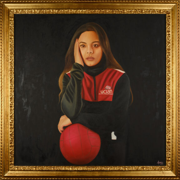 Image of a person in a red and black UCLan zipper jacket leaning on a sports ball.