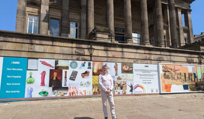 A person wearing a white shirt and beige trousers stands in front of The Harris on a sunny day.