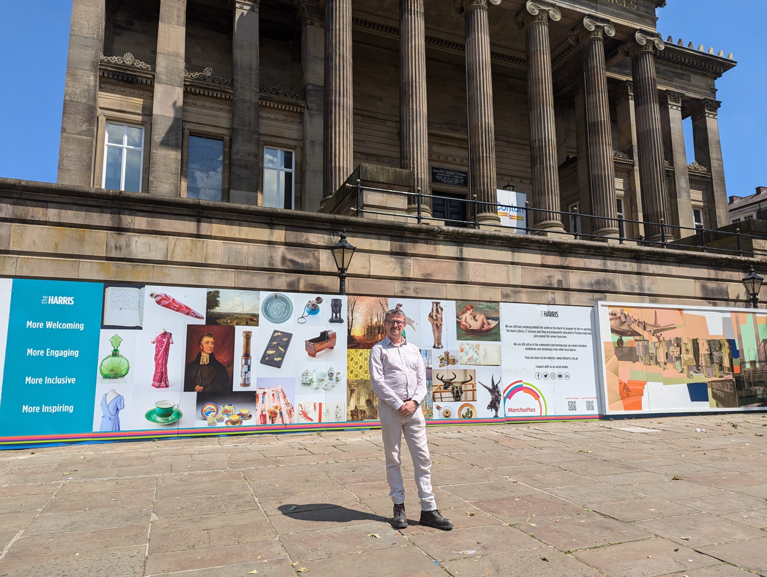 A person wearing a white shirt and beige trousers stands in front of The Harris on a sunny day.