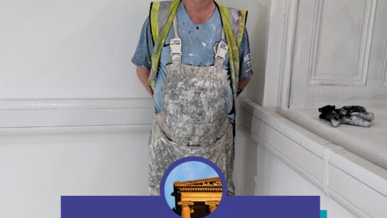 A person wearing overalls, hard hat and high vis stands in front of a freshly painted wall.