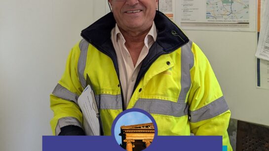 A person wearing a hard hat and protective clothing stands in an office.