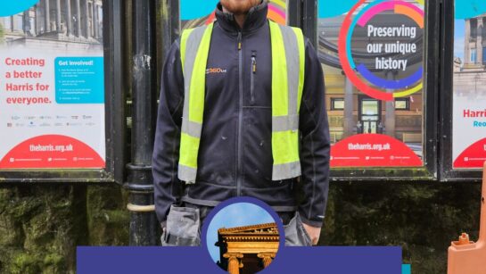 A person wearing a hard hat and protective clothing stands in front of colourful posters.