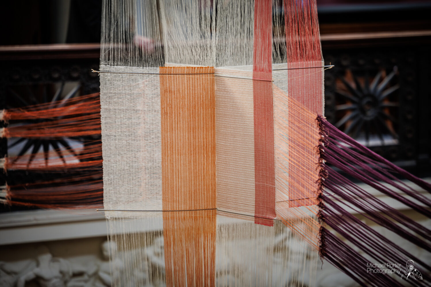 Close-up of colorful weaving threads, including white, orange, and maroon, interlaced in a geometric pattern. The background is blurred, highlighting the intricate textile art.