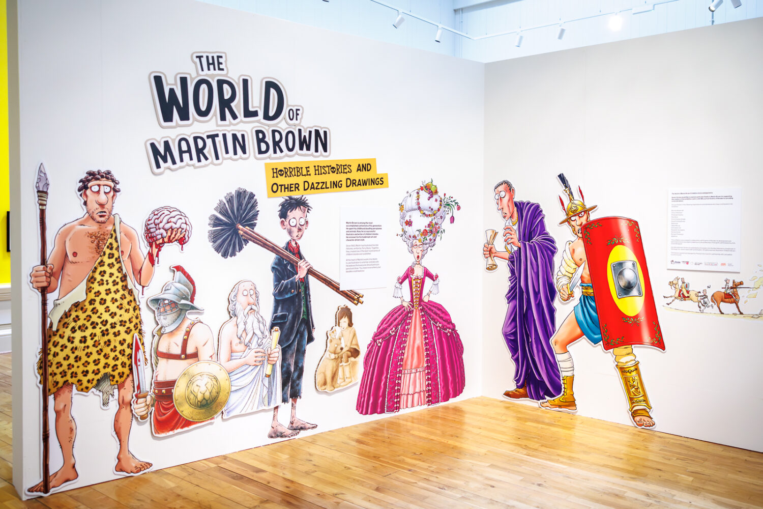 Exhibit wall showcasing Martin Brown's humorous historical illustrations from "Horrible Histories," featuring cartoonish, colorful historical figures.