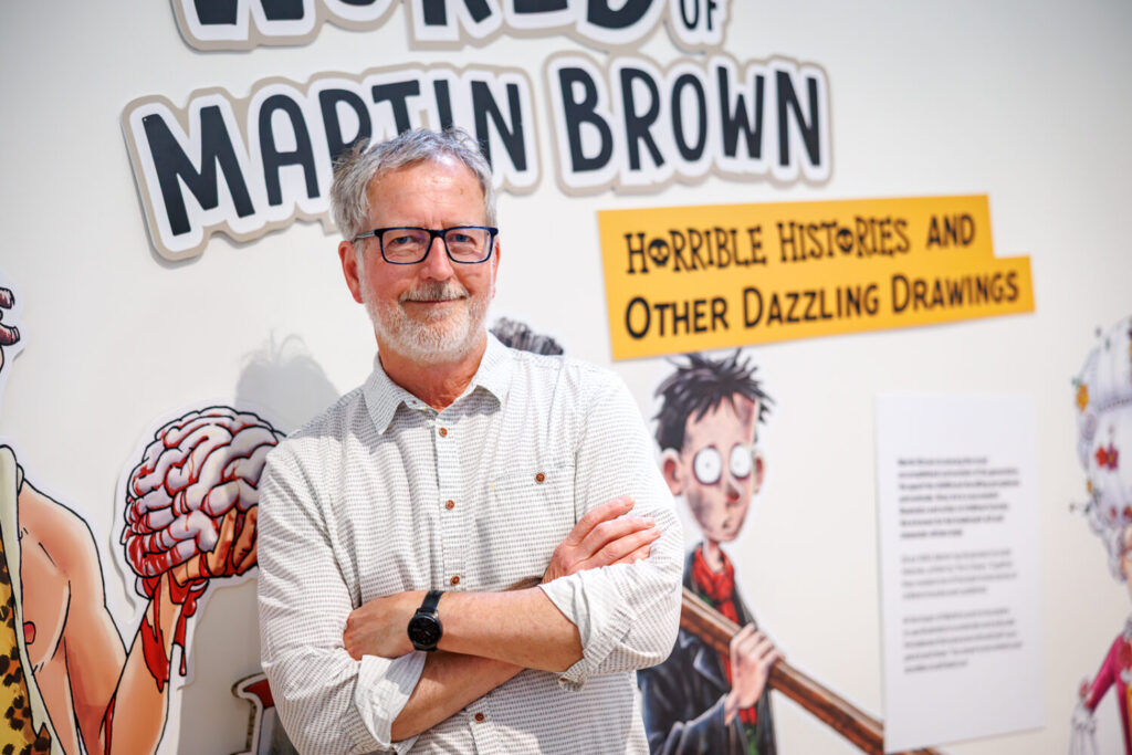 Exhibit wall showcasing Martin Brown's humorous historical illustrations from "Horrible Histories," featuring cartoonish, colorful historical figures.