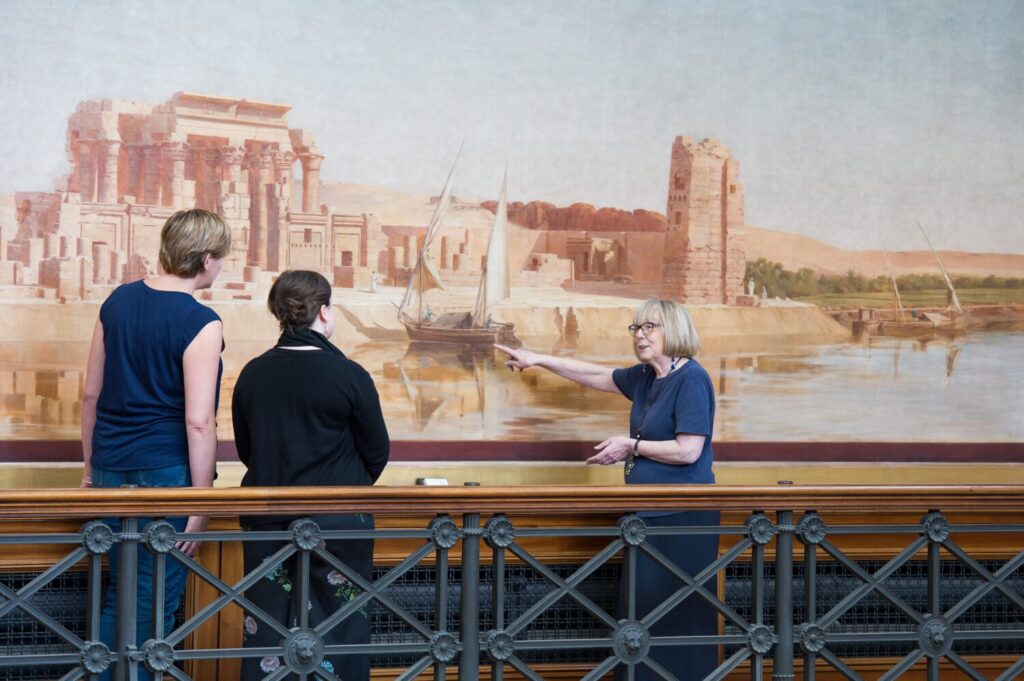 A guide points towards a large painting of Ancient Egypt whilst two people watch.