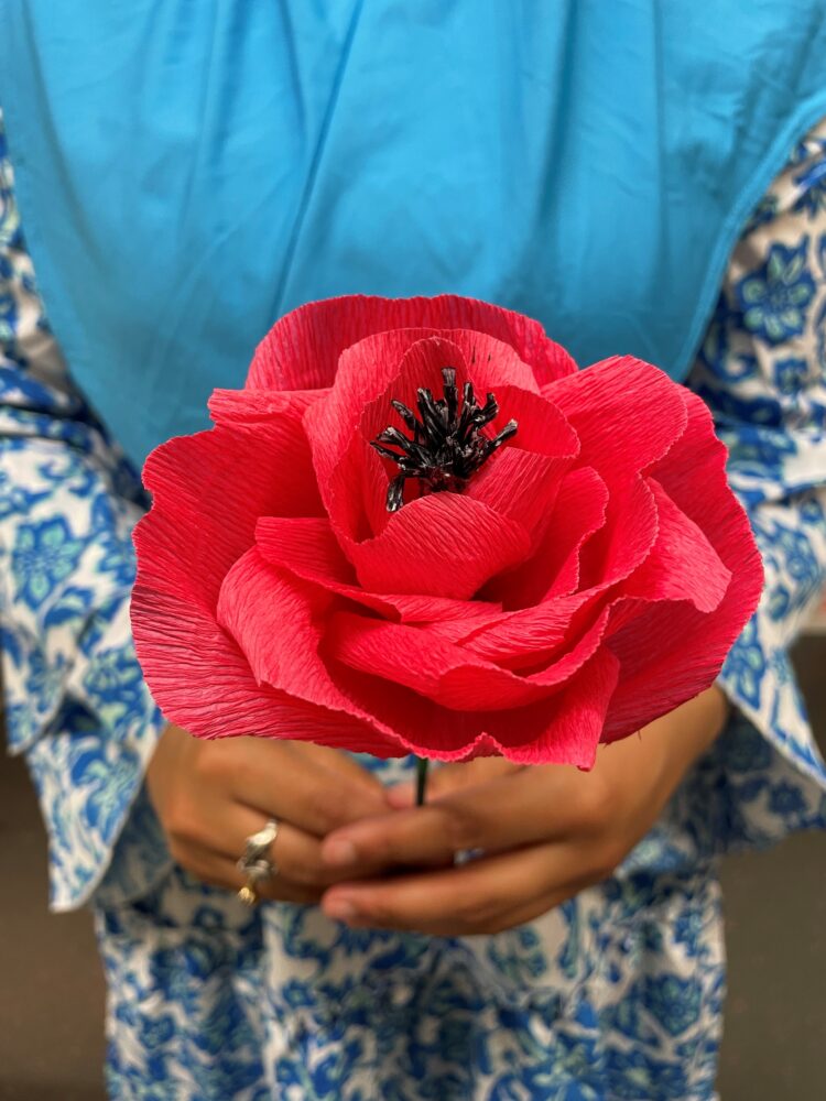 A brown ceramic jug with a blue handle holds a twisted branch with three white paper flowers, featuring vibrant red centers. Set against a plain beige wall, the arrangement has a minimalist and delicate aesthetic.