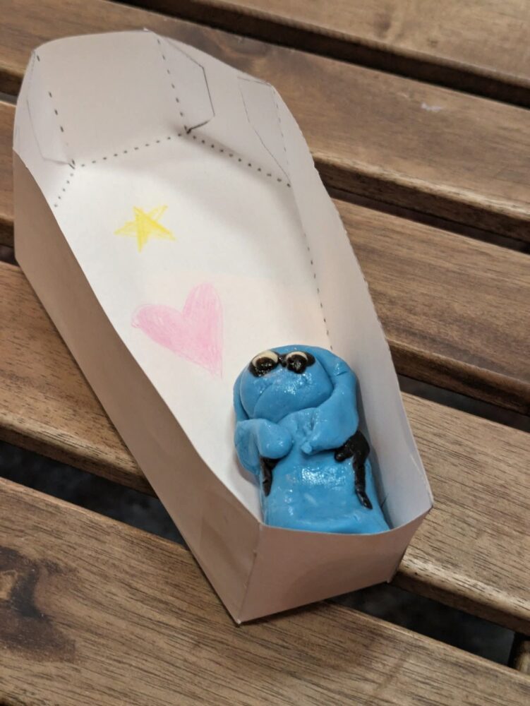 A small blue figurine is lying in a white paper coffin with a hand-drawn pink heart and yellow star. It's on a wooden surface, conveying a whimsical tone.