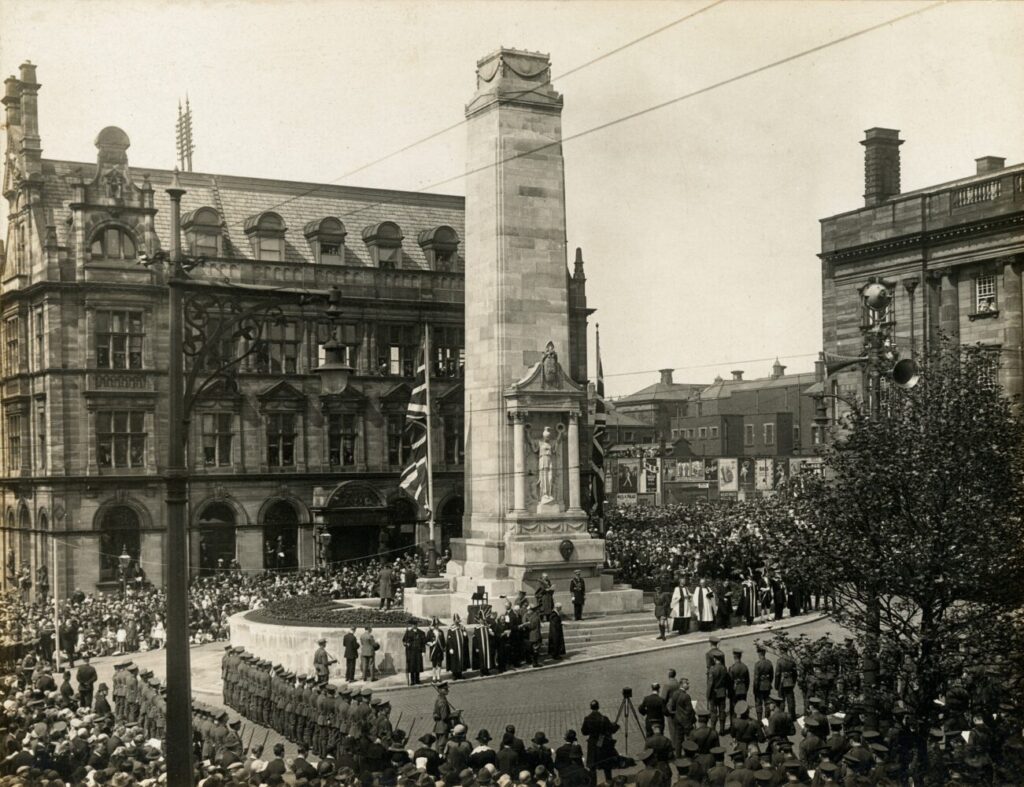 Historic photograph of a large crowd gathered around a tall war memorial in an urban square. Buildings surround the monument, with flags and somber atmosphere.