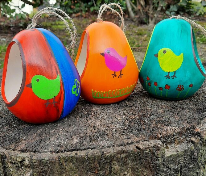 Three colorful, hand-painted gourds with bird designs sit on a tree stump. The gourds, orange, blue, and green, have twine loops for hanging.
