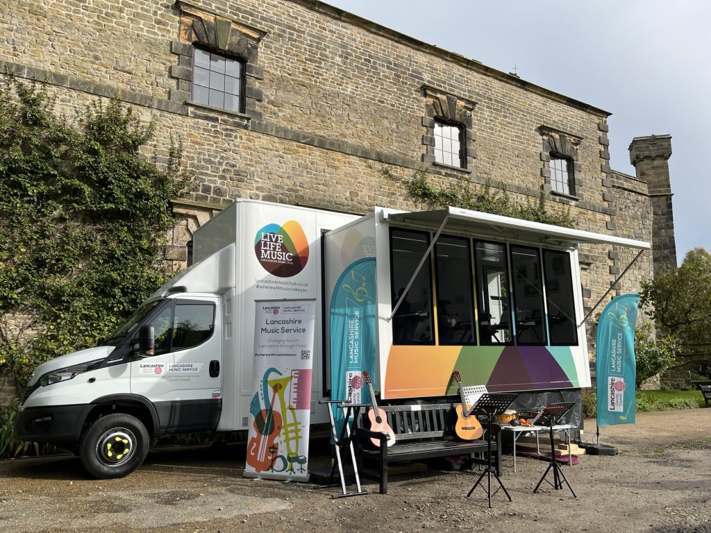 A colorful mobile music service truck with "Live Life Music" branding is parked beside an old brick building. Guitars and music stands are set up outside.