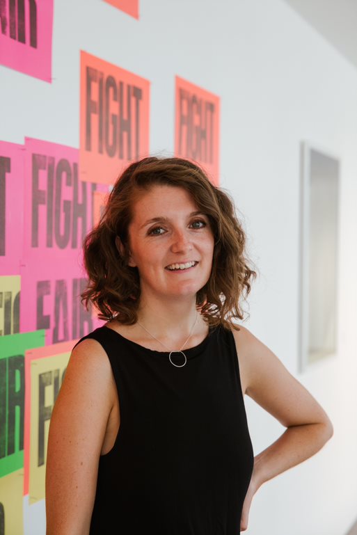 A woman with curly hair smiles confidently in front of a wall with colorful posters reading "FIGHT." She exudes a sense of empowerment and positivity.
