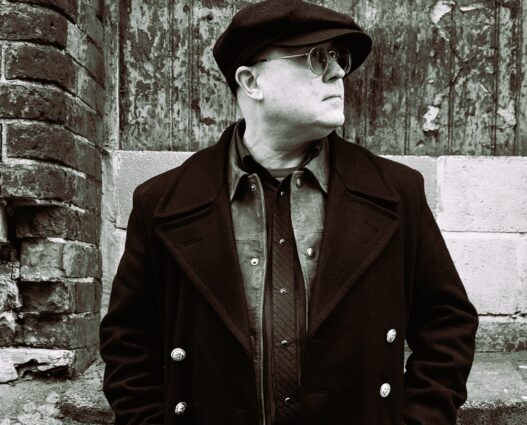 A person in vintage attire leans against a brick wall, wearing a long coat, flat cap, and sunglasses. The setting and style evoke a classic, timeless feel.