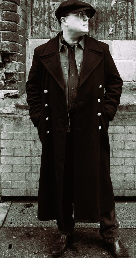 A person in vintage attire leans against a brick wall, wearing a long coat, flat cap, and sunglasses. The setting and style evoke a classic, timeless feel.