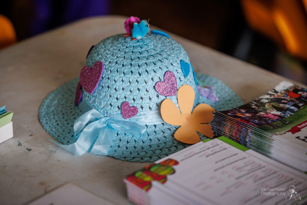 A blue woven hat decorated with glittery heart shapes and a paper flower sits on a table next to colorful brochures, evoking a playful and creative vibe.