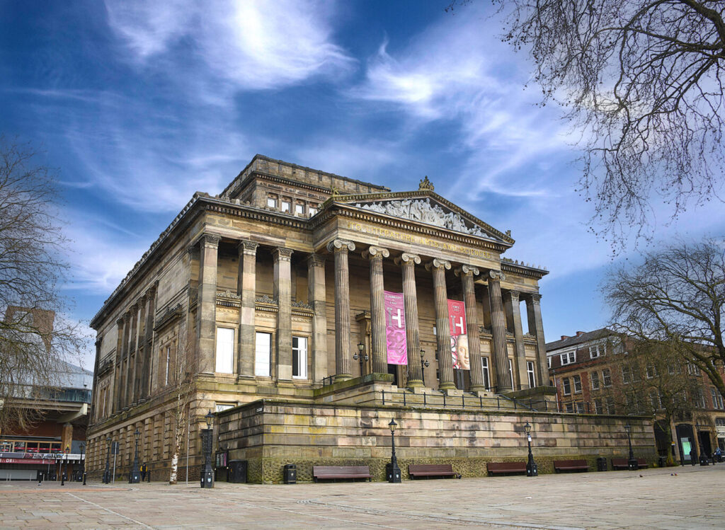 Majestic neoclassical building with tall columns and intricate carvings, set against a vibrant blue sky. Pink banners hang, adding a touch of modernity.
