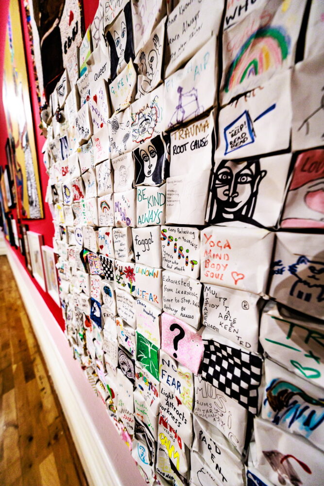 A colorful wall collage of small, creatively decorated squares with drawings and words. The vibrant and varied designs evoke a sense of community and expression.