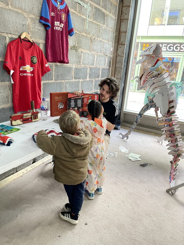 A person in a "Team Member" shirt shows two children a display table with historical items. Soccer jerseys hang on a brick wall. The scene feels educational and interactive.
