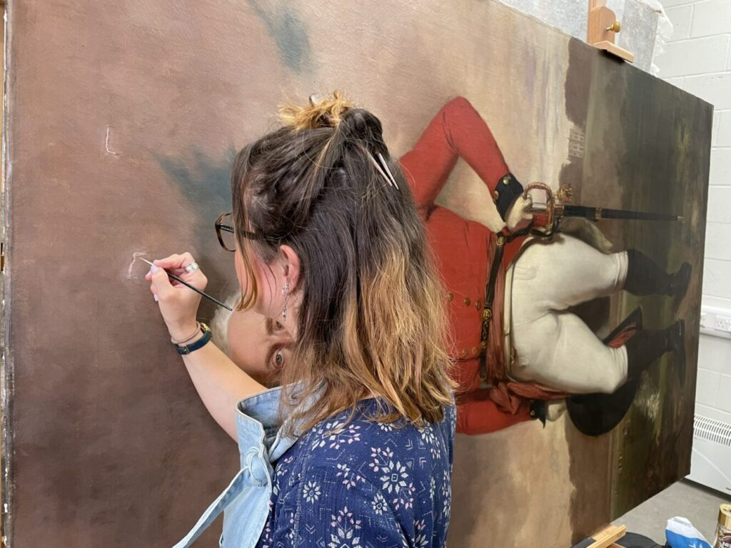 A woman carefully restores a large painting, focusing intently. The artwork depicts a soldier in a red coat, evoking a sense of historical preservation.