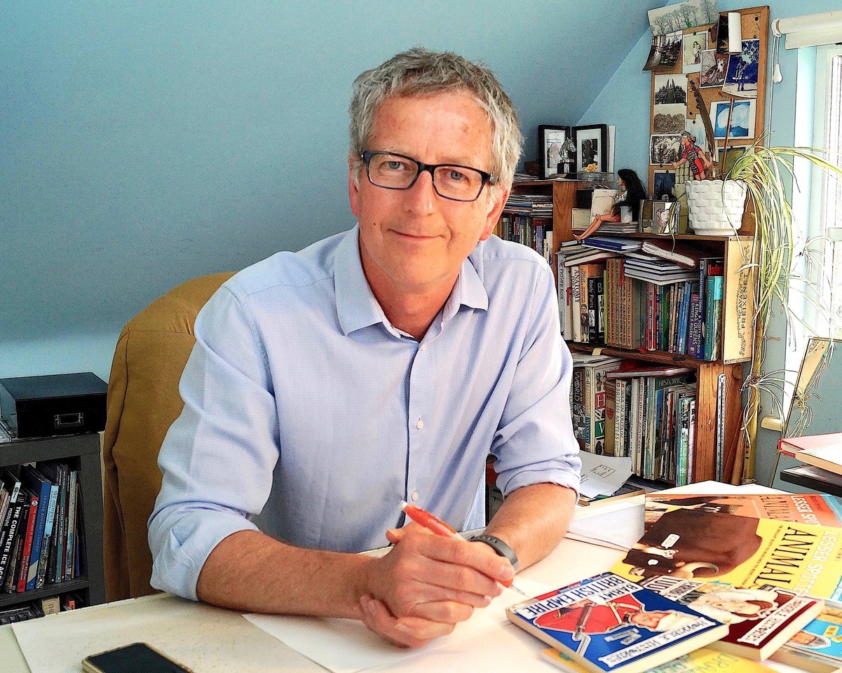 Elderly man with glasses, wearing a blue shirt, sits at a cluttered desk with books and magazines. Shelves and photos fill the background, creating a cozy, intellectual setting.
