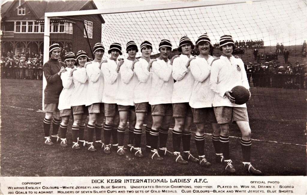 Black and white photo of a historic women's soccer team wearing white jerseys, blue shorts, and striped hats, standing in a line on a field, smiling confidently.