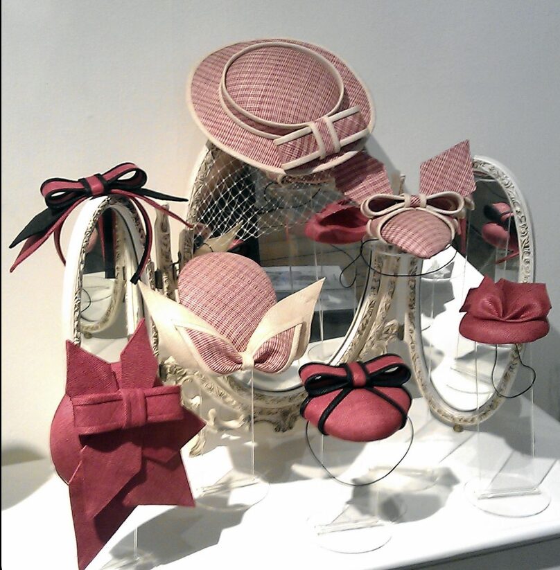 Elegant display of various red and pink hats with bows and ribbons, set against ornate mirrors. The scene conveys a vintage and stylish tone.