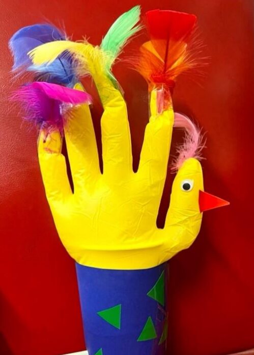 A colorful hand-shaped paper craft resembling a bird, with googly eyes, red beak, and vibrant feathers, set against a red background.