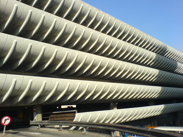 Concrete parking structure with distinctive curved, ribbed surfaces creates a rhythmic pattern. A road curves under a bright blue sky.