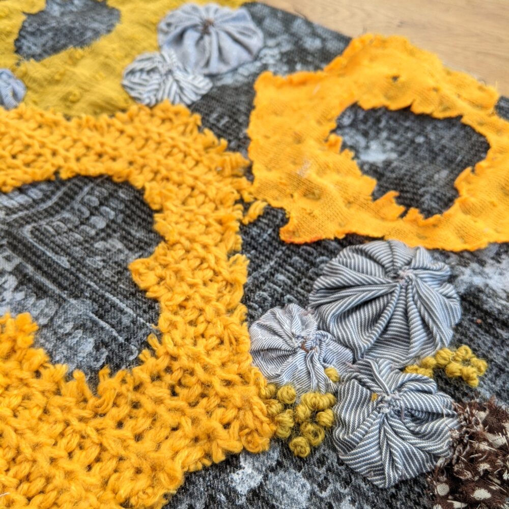 Close-up of a patchwork textile featuring vibrant yellow crochet circles and textured fabric flowers on a gray patterned background, evoking a crafted, warm feel.