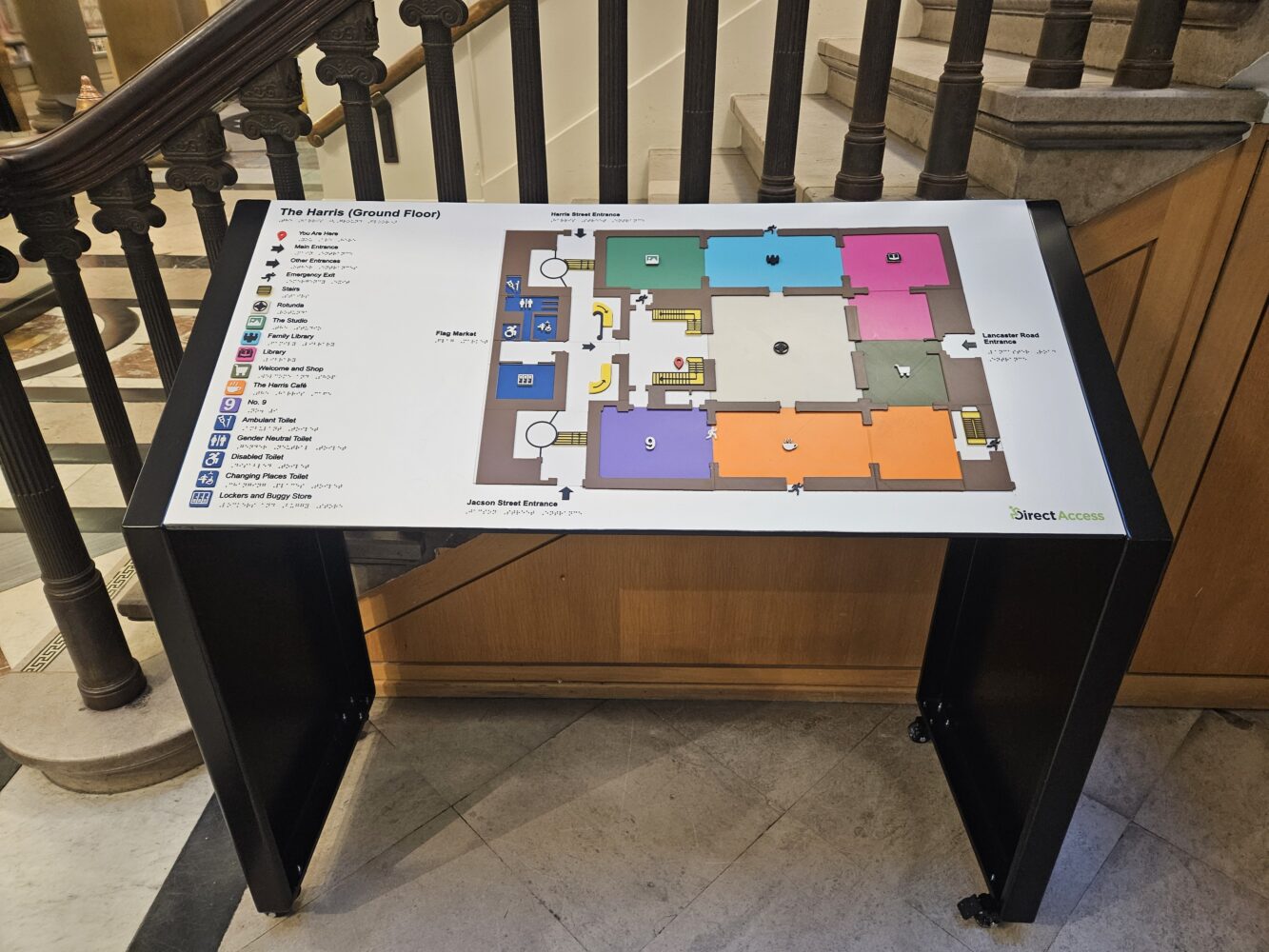 Museum interior with a colorful floor map displayed on a stand near a staircase. Visitors gather in the background, creating a lively atmosphere.