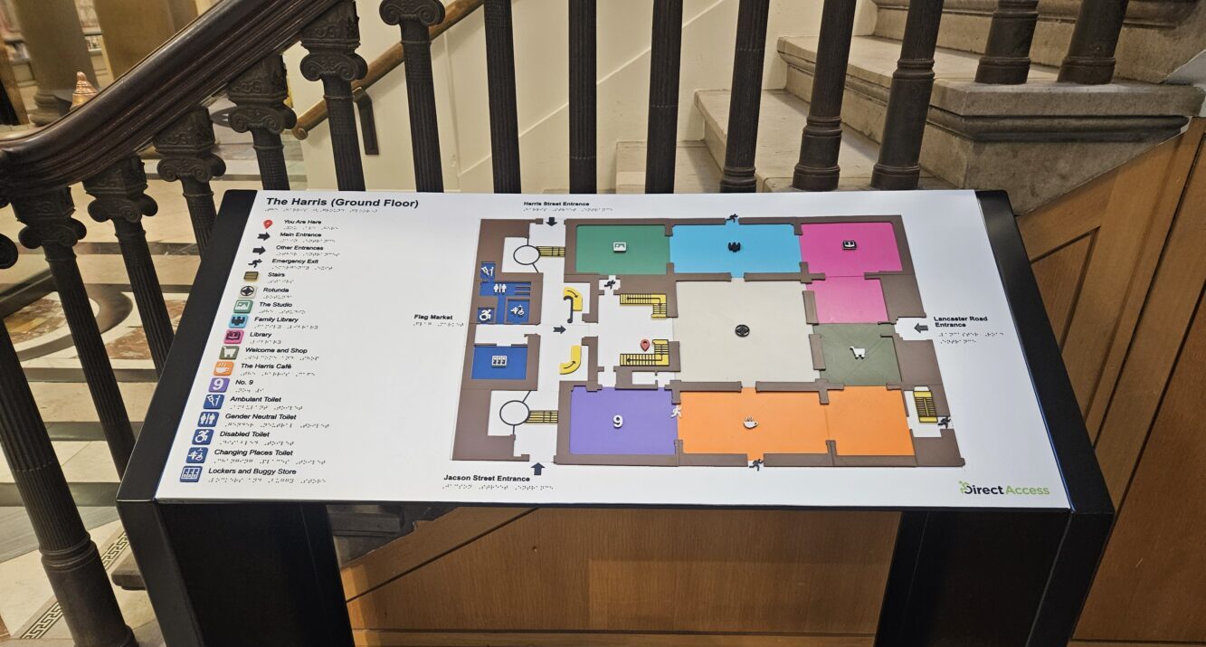 Museum interior with a colorful floor map displayed on a stand near a staircase. Visitors gather in the background, creating a lively atmosphere.