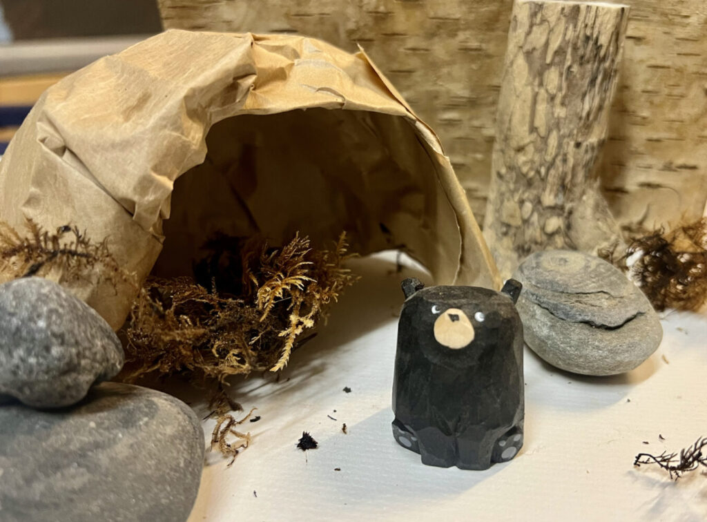 A small, black bear figurine sits in front of a rustic woodland scene. The bear is near a paper cave with moss, surrounded by stones and logs. Cozy and natural atmosphere.