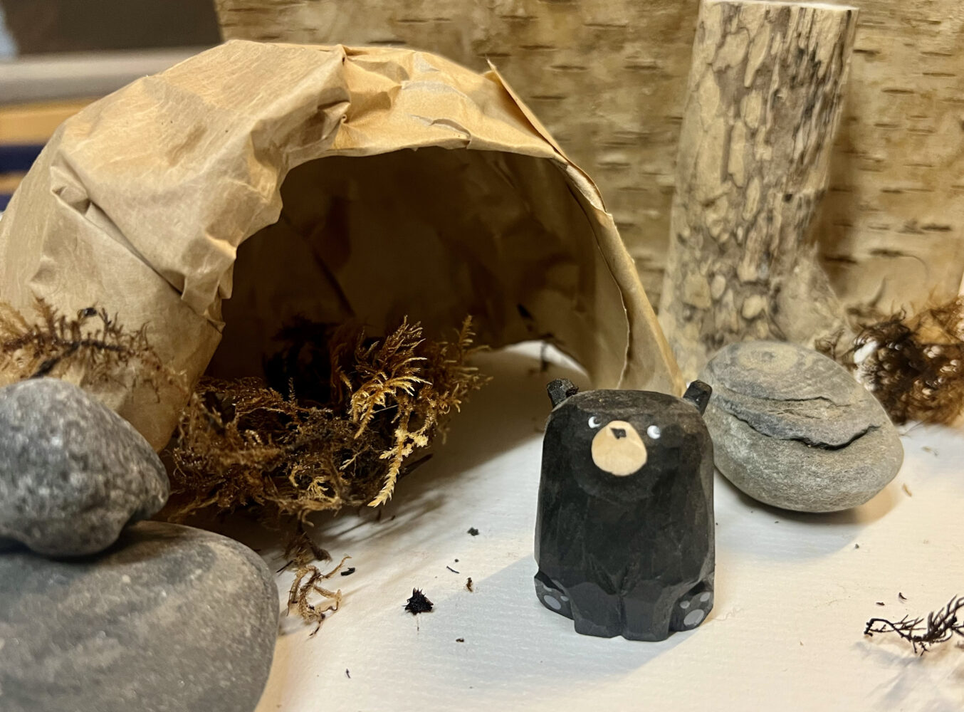 A small, black bear figurine sits in front of a rustic woodland scene. The bear is near a paper cave with moss, surrounded by stones and logs. Cozy and natural atmosphere.
