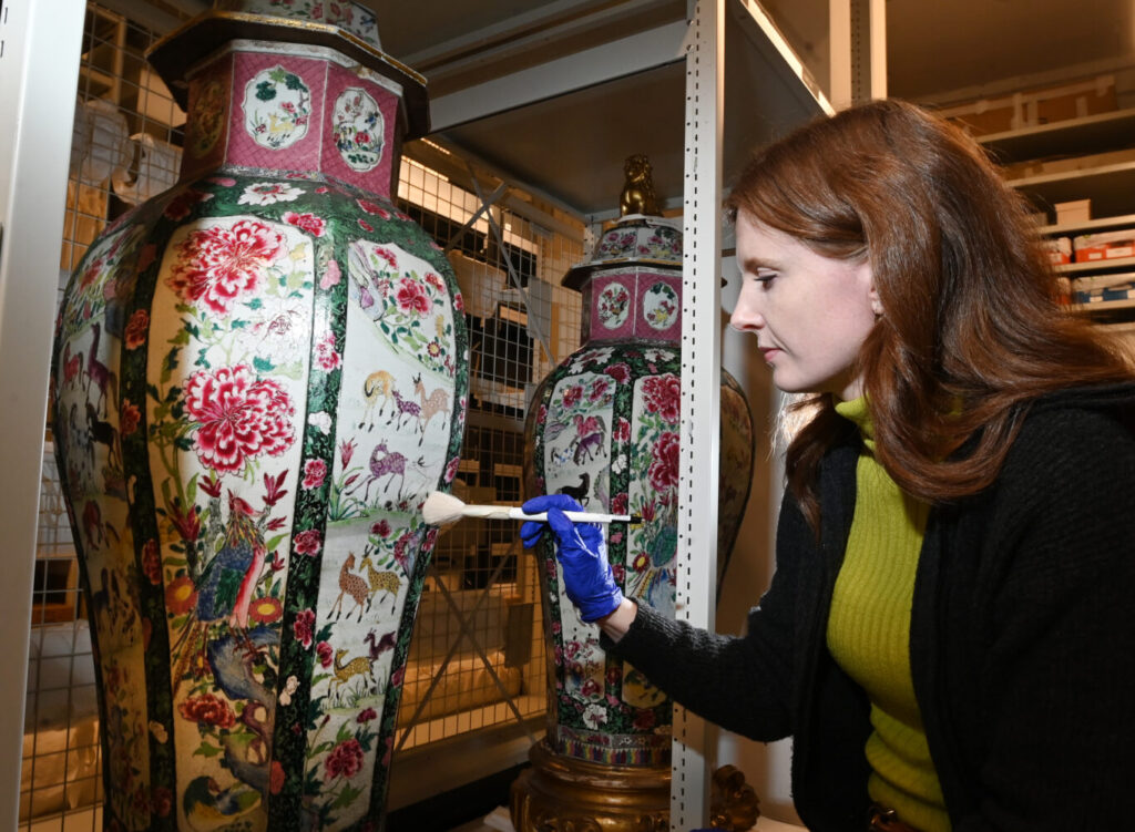 A person with red hair carefully dusts a large, ornate vase featuring colorful floral and animal designs. They wear a yellow sweater and blue gloves, conveying focus and care. The setting is a storage or museum area with shelves.