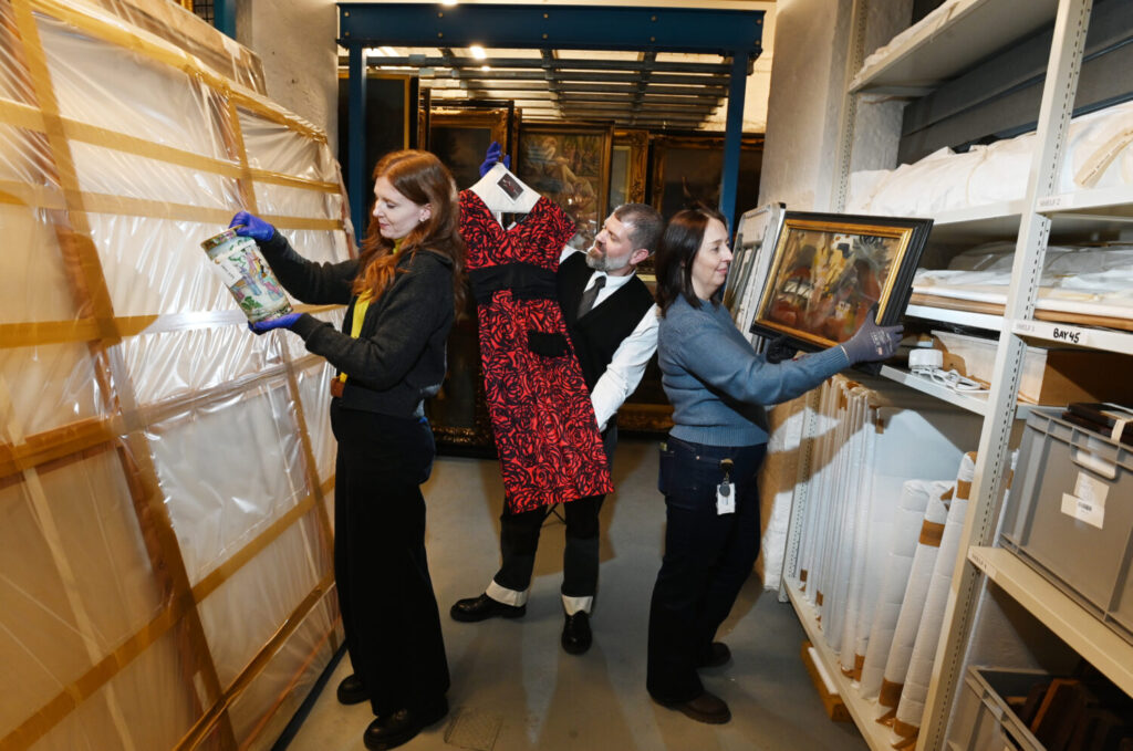 Three people carefully handle items in an art storage room. They examine a colorful vase, a red patterned dress, and a framed painting, conveying a sense of careful preservation.