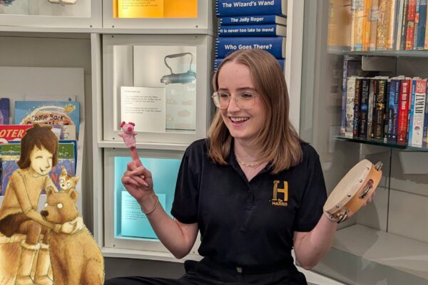 A woman in a museum setting smiles while holding a pink puppet and tambourine. She's beside an illustration of a child with a kangaroo, surrounded by books.