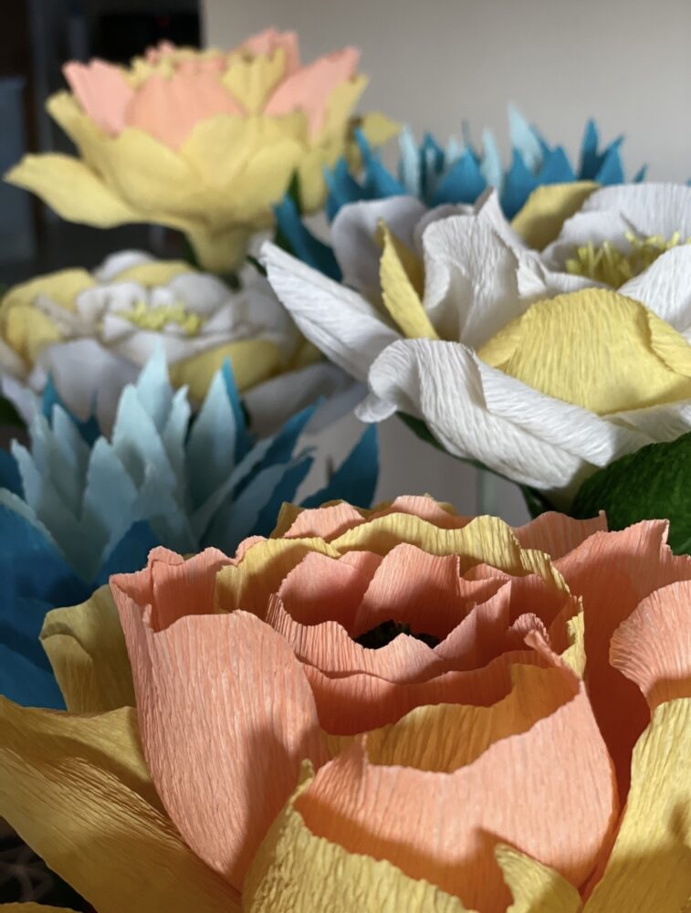 Close-up of colorful paper flowers with intricate petals in peach, yellow, white, and blue. The arrangement feels vibrant and artistic.