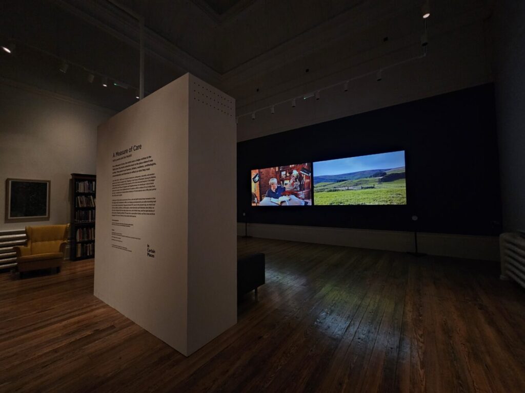 Dimly lit gallery with a display titled "A Measure of Care." Two large screens show a person reading and a serene landscape. Warm, contemplative atmosphere.