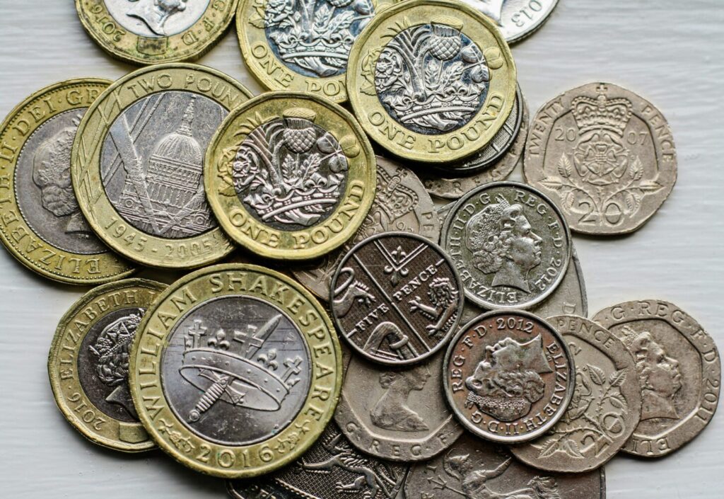 A collection of British coins, including one and two-pound coins with distinct designs, lies scattered on a surface. The coins are metallic with various patterns.