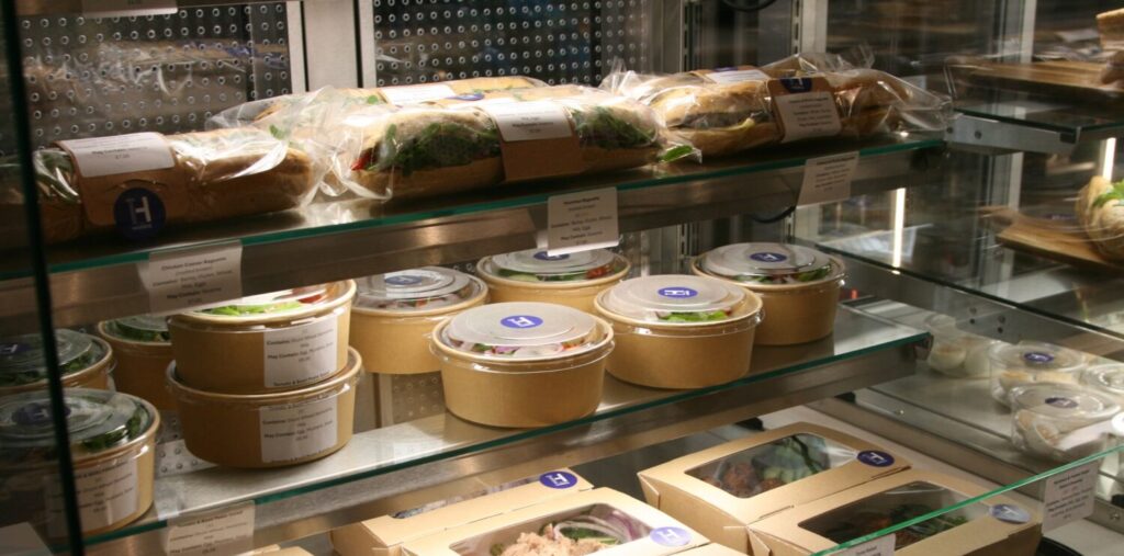 Display case with packaged food: sandwiches, salads, and boxed meals. Items are neatly arranged with labels, offering a fresh, convenient selection.