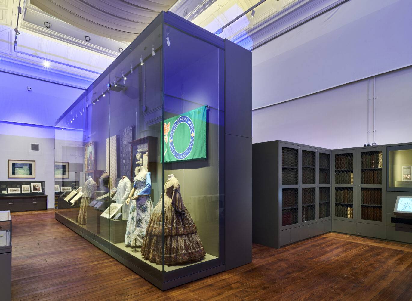 A museum exhibit displays vintage dresses in a large glass case. A colorful flag hangs above. The room is softly lit with wooden floors, creating a historical ambiance.