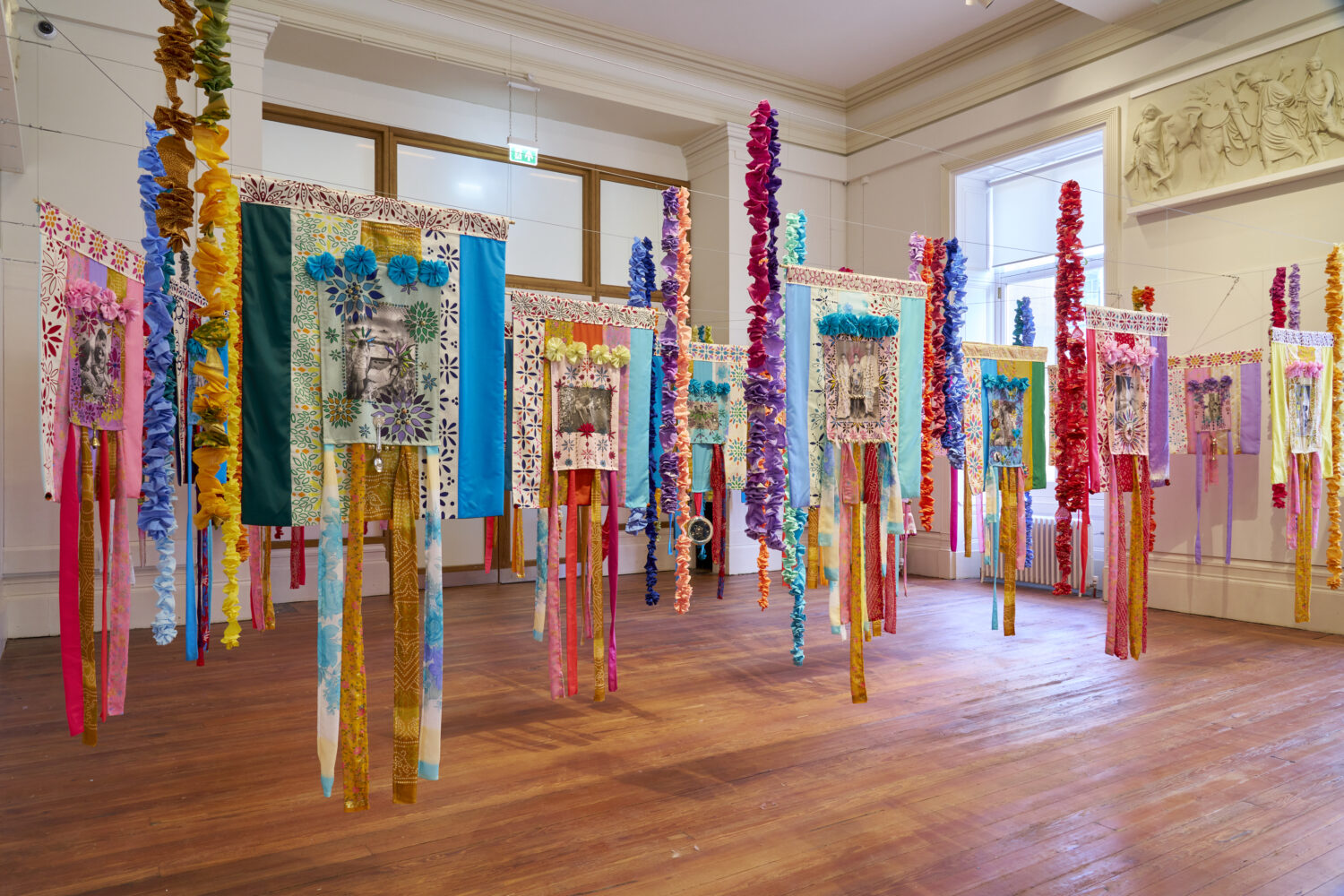 Vibrant textile banners with floral patterns and colorful ribbons hang in a bright gallery, conveying a festive and lively atmosphere.