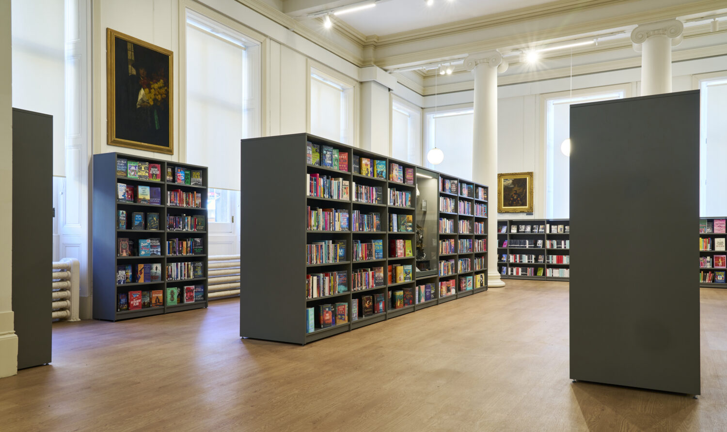 A spacious, well-lit library interior with tall, white columns, wooden floors, and shelves filled with colorful books. Walls have paintings and large windows.