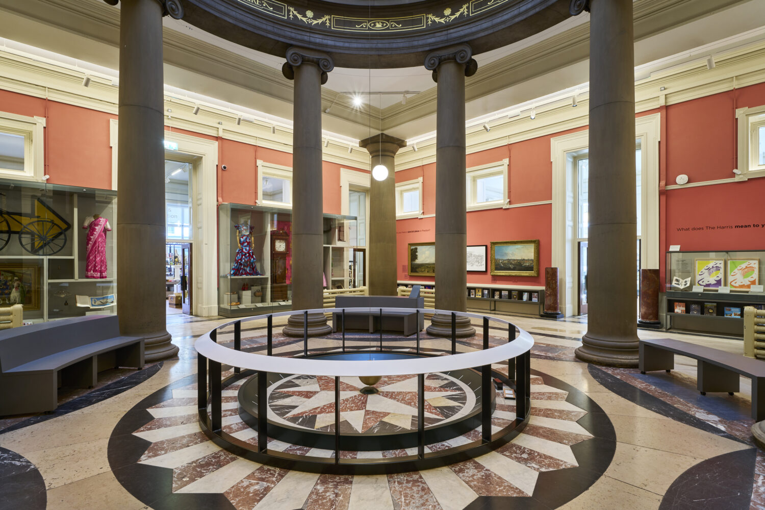 Spacious museum interior with circular layout, red walls, and tall columns. Exhibits include colorful art and dresses, creating an inviting atmosphere.